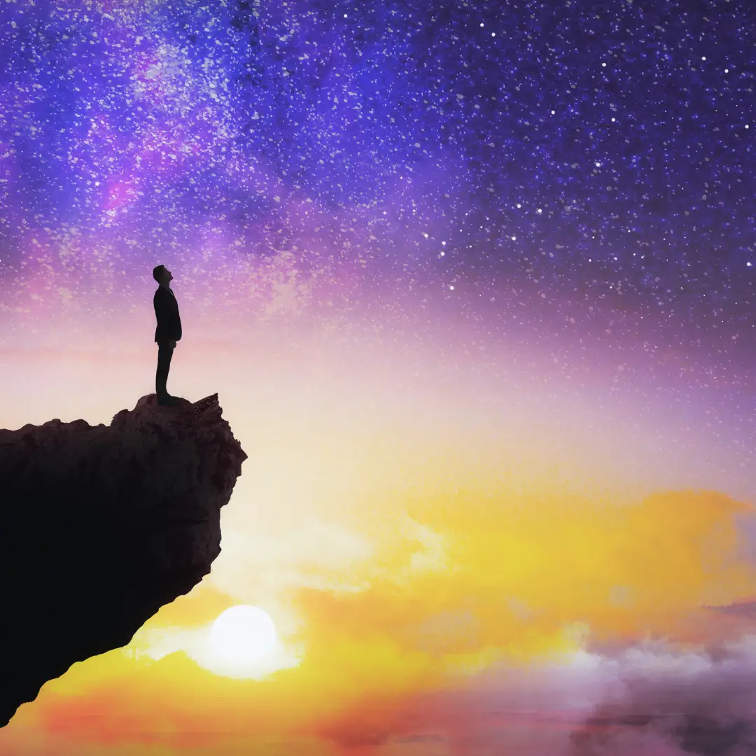 person standing on edge of cliff staring out at purple and orange sunset sky with stars, contemplating life and consciousness